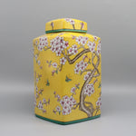 Un pot de stockage vintage décoré de motifs floraux élégants en fleurs de cerisier sur un fond jaune vif, avec de délicats papillons volant autour.