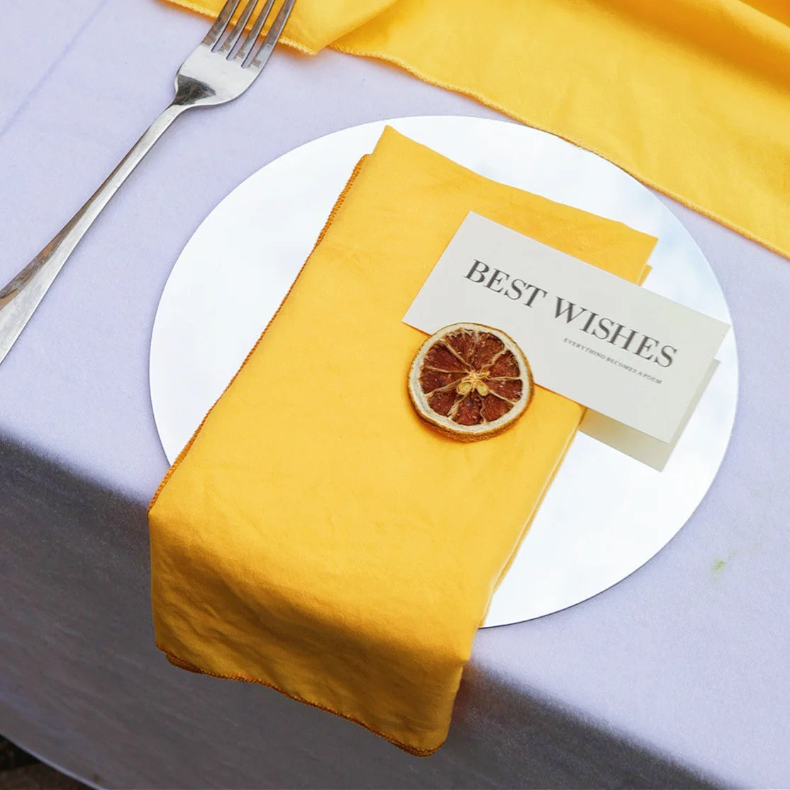 Une nappe et une serviette de table jaunes accompagnent une assiette blanche contenant un carton indiquant "BEST WISHES" ainsi qu'une tranche d'orange séchée comme décoration.