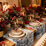 La table de Noël est magnifiquement décorée avec des guirlandes de sapins, des baies rouges, des bougies allumées et des accessoires de fête, créant une ambiance chaleureuse et festive.