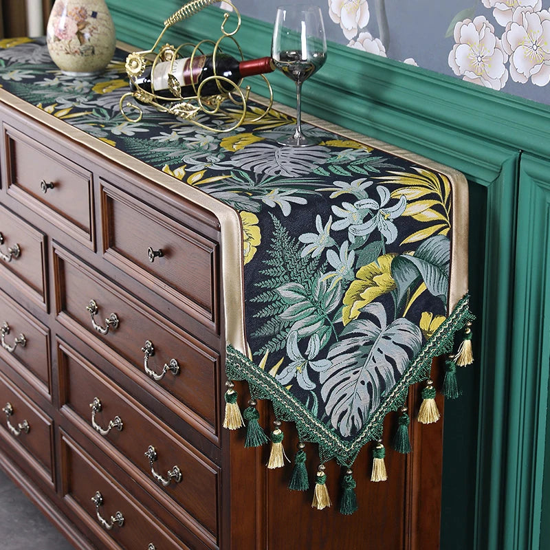 Cette image montre une commode de luxe en bois foncé sur laquelle est posée une nappe ornée de motifs floraux tropicaux aux couleurs chatoyantes, rehaussée de pompons verts.