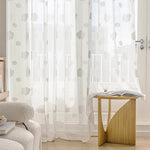 Rideau voilage blanc à motifs de nuages dans un salon lumineux, avec une table en bois sculpté et un fauteuil bouclette crème.