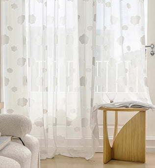 Rideau voilage blanc à motifs de nuages dans un salon lumineux, avec une table en bois sculpté et un fauteuil bouclette crème.