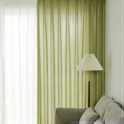 Grand rideau vert amande habillant une baie vitrée, avec un canapé gris et un lampadaire blanc.