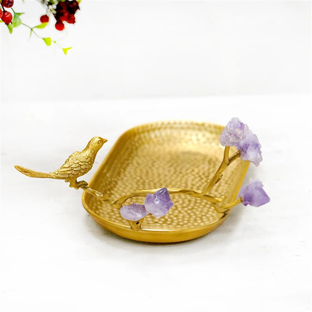 Un bol décoratif en métal doré avec un oiseau sculpté et des cristaux d'améthyste, sur fond de fleurs rouges et vertes.