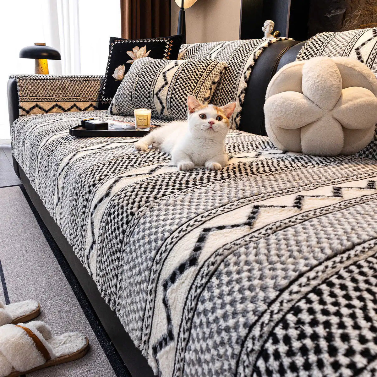 L'image montre un chat blanc et beige assis confortablement sur un canapé recouvert d'un motif géométrique noir et blanc, avec de nombreux coussins et décorations dans le même style.