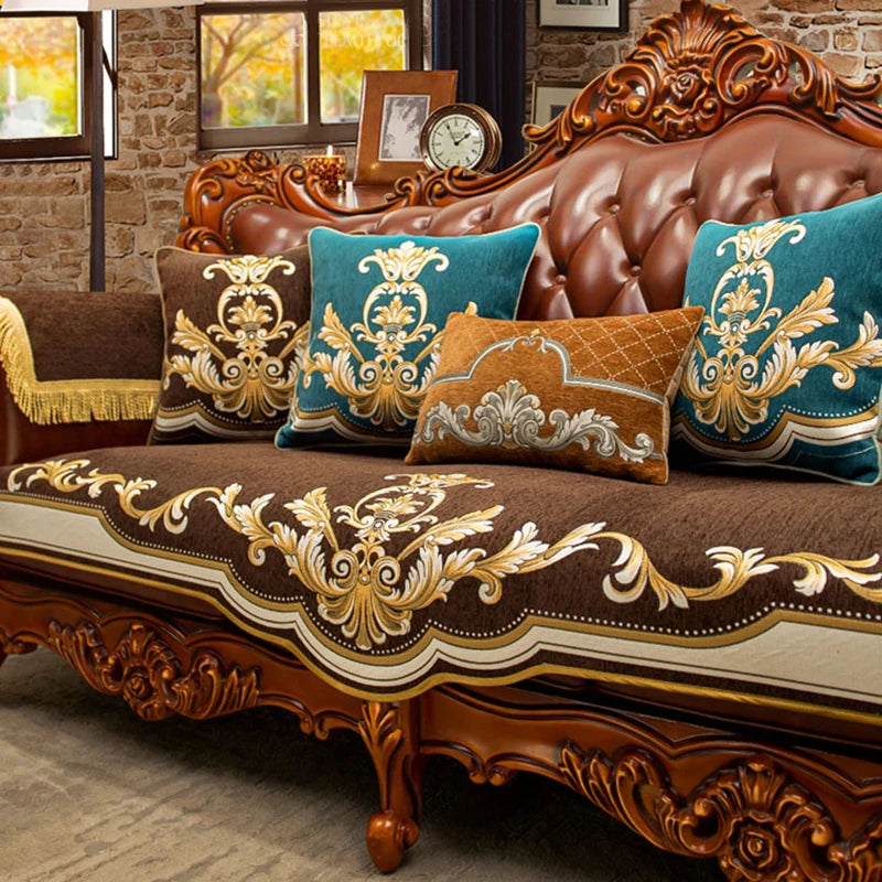 Élégant canapé en cuir marron foncé orné de motifs dorés baroque et de coussins décoratifs bleu-vert et dorés, dans un cadre rustique avec fenêtres offrant une vue sur un paysage automnal.