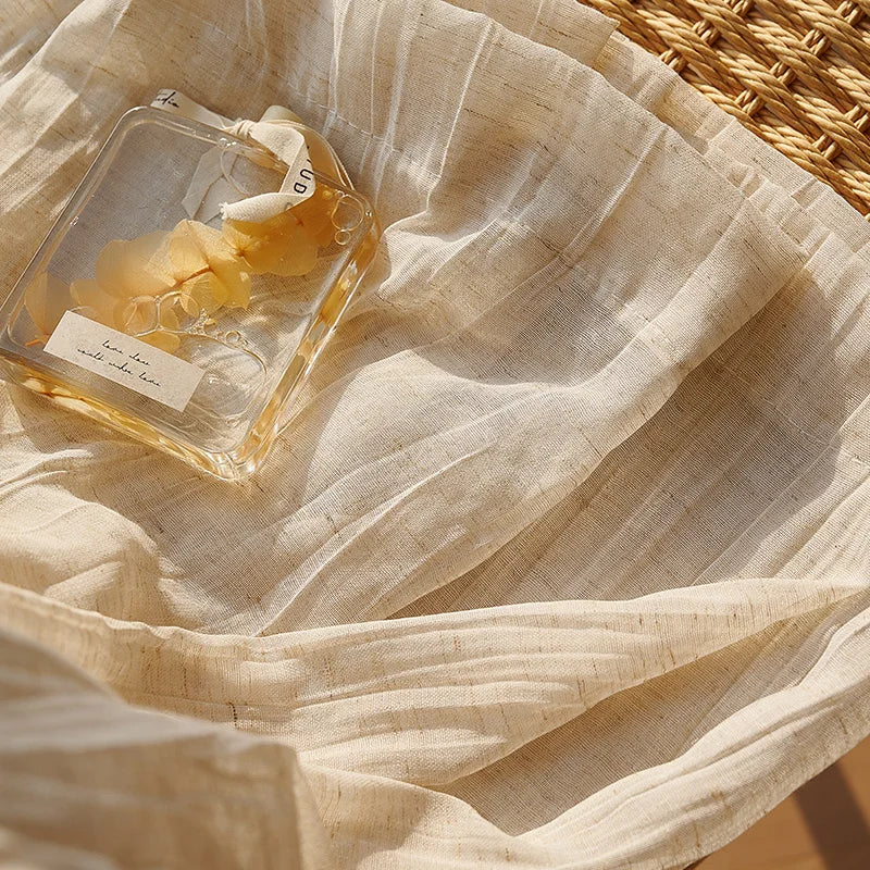 Gros plan sur un voilage d'été beige clair, légèrement froissé, baigné de lumière naturelle, avec un flacon de parfum en verre posé délicatement dessus.