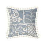 Coussin décoratif gris clair aux motifs floraux et géométriques, avec des pompons blancs sur les bords.