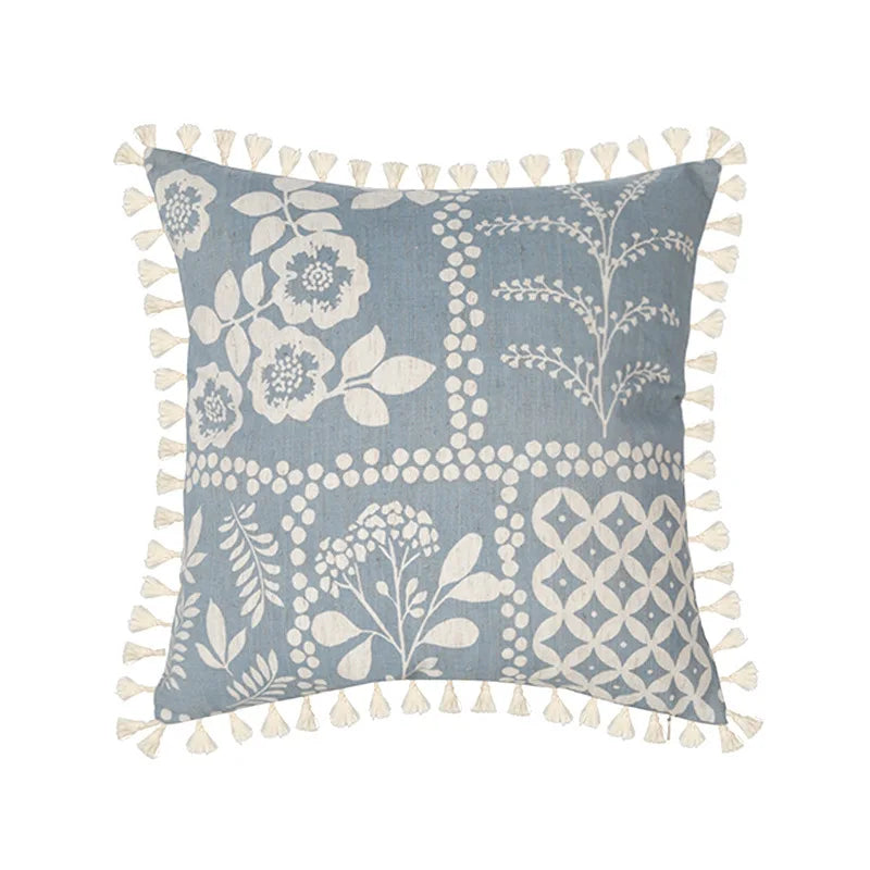 Coussin décoratif gris clair aux motifs floraux et géométriques, avec des pompons blancs sur les bords.