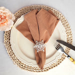 Un ensemble de table décoré avec un napperon tressé, une assiette blanche, des serviettes marron et un ornement en métal en forme de flocon de neige, accompagné d'un bouquet de fleurs roses.