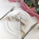 Une nappe blanche avec une serviette de table grise ornée de motifs floraux et de plantes vertes, accompagnée d'une fourchette et d'un verre de vin sur la table.