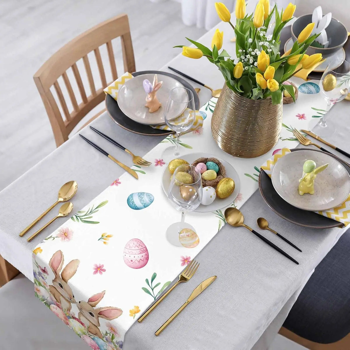 Cette table dressée pour Pâques présente de belles décorations printanières, notamment un bouquet de tulipes jaunes dans un vase doré, des assiettes noires et dorées, des couverts dorés et des oeufs de Pâques colorés.
