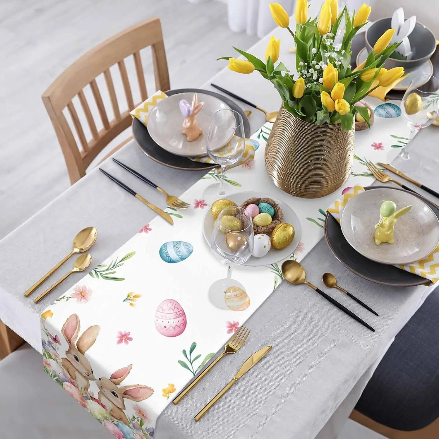 Cette table dressée pour Pâques présente de belles décorations printanières, notamment un bouquet de tulipes jaunes dans un vase doré, des assiettes noires et dorées, des couverts dorés et des oeufs de Pâques colorés.