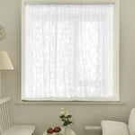 Voilage blanc court à motifs floraux, habillant une petite fenêtre dans un intérieur lumineux.