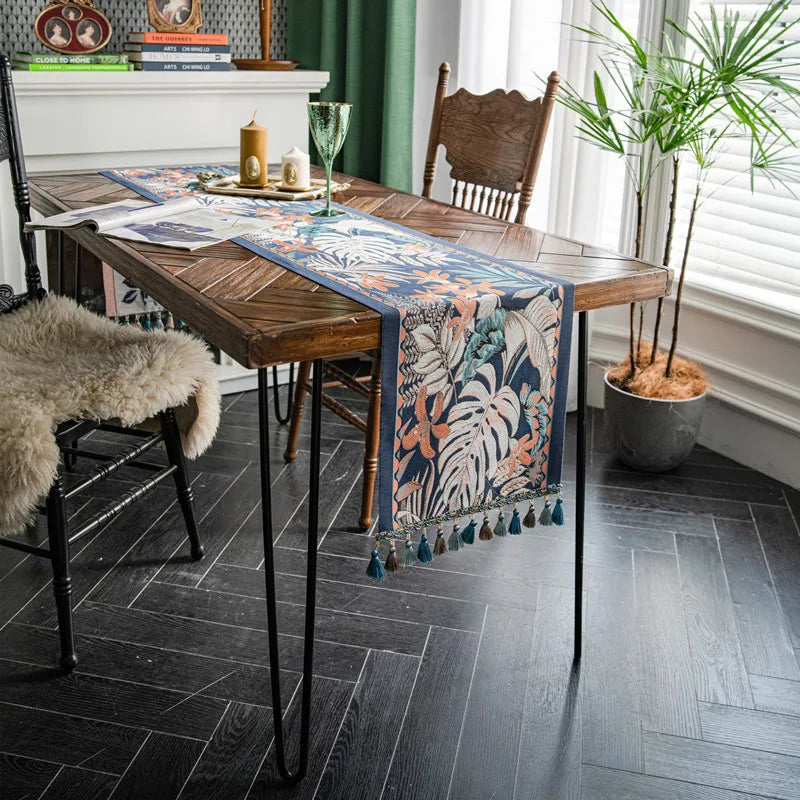 La photo montre une table en bois massif avec un chemin de table orné de motifs floraux et de pompons, sur un sol en ardoise, entourée de plantes et d'éléments de décoration rustiques.