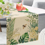 Cette image montre un beau chemin de table beige orné de motifs de feuilles tropicales vertes, de fleurs jaunes et d'une abeille, créant une ambiance naturelle et chaleureuse.