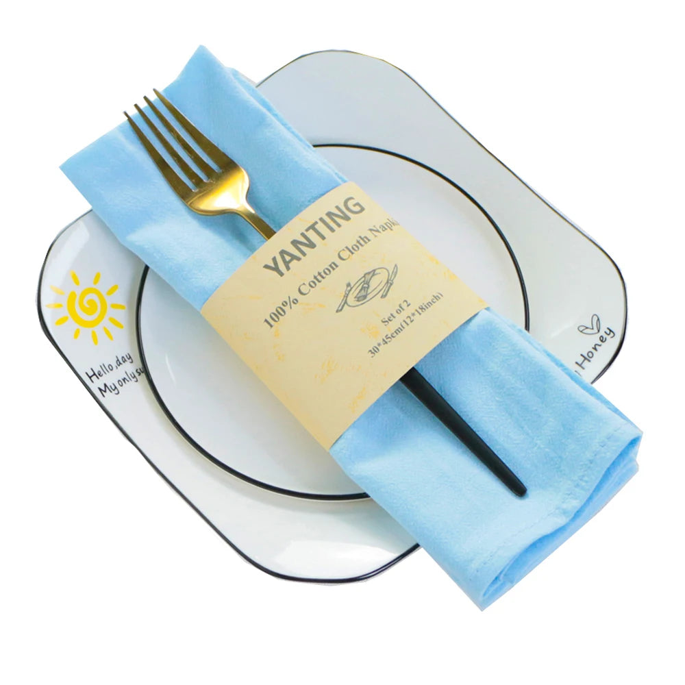 Sur une assiette blanche se trouve une serviette en tissu bleu ciel enroulée autour d'une fourchette dorée, avec un emballage jaune sur lequel est inscrit "Wanting 100% Cotton Cloth Napkin".