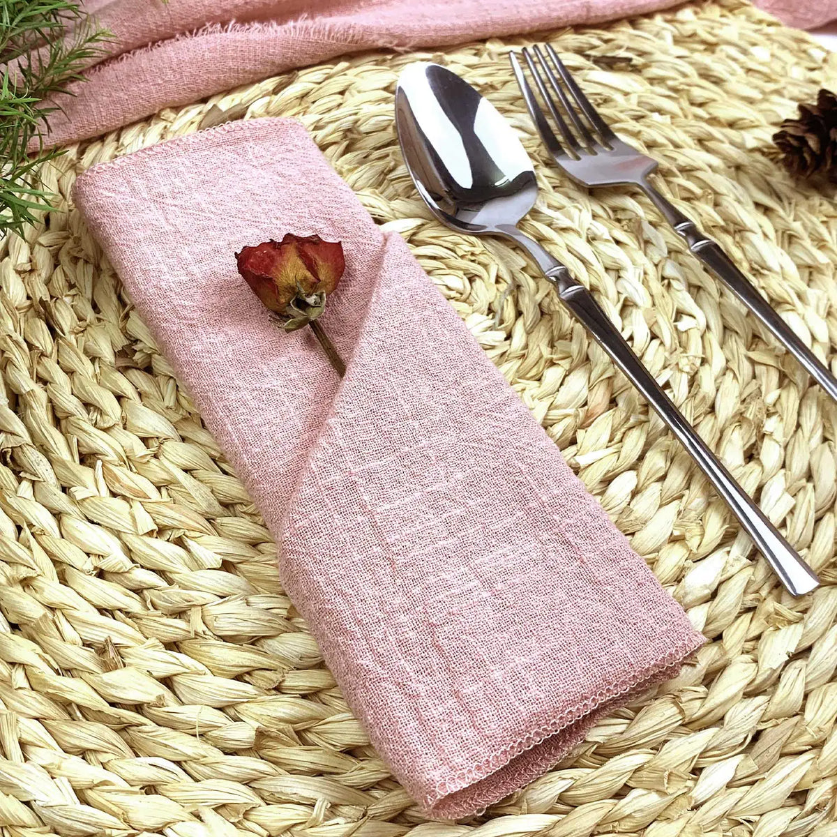 Réglage de table élégant avec une serviette rose pastel et une fleur séchée, posé sur un tapis en fibres tressées.