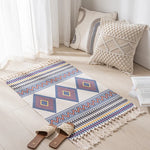 Tapis décoratif avec des motifs géométriques colorés et des franges, posé sur le sol d'un intérieur lumineux avec des coussins et des rideaux blancs.