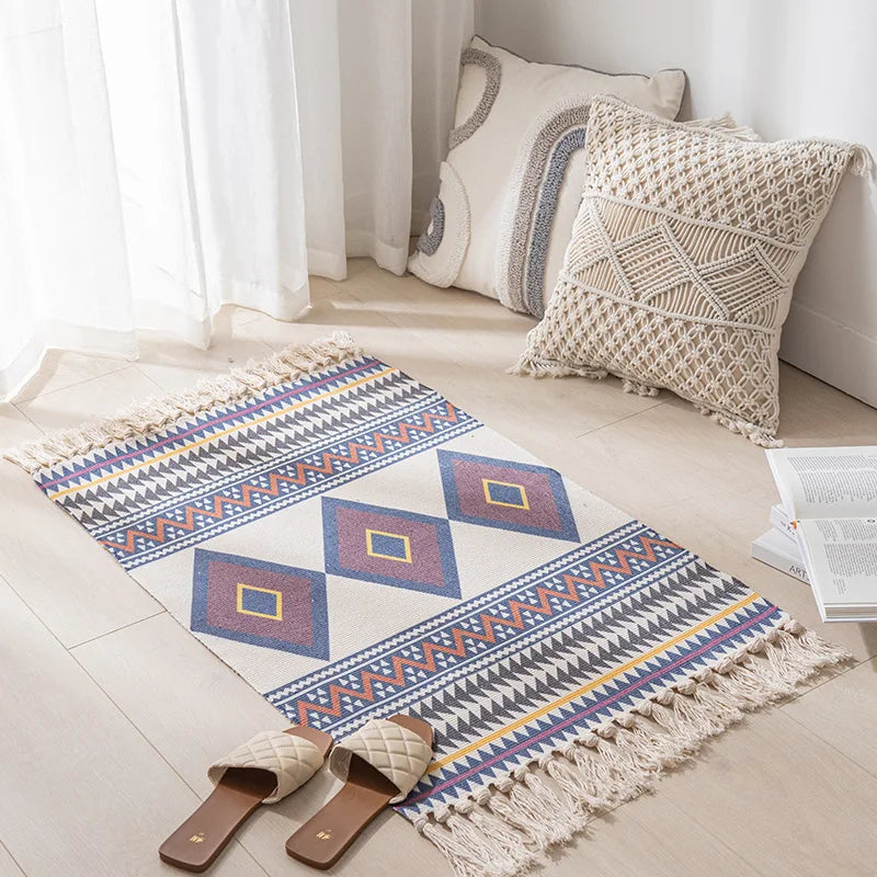 Tapis décoratif avec des motifs géométriques colorés et des franges, posé sur le sol d'un intérieur lumineux avec des coussins et des rideaux blancs.