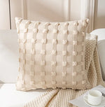 Coussin décoratif texturé avec des motifs carrés et pompons blancs, posé sur un canapé blanc dans un intérieur lumineux.