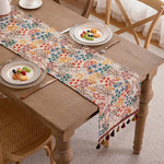 Chemin de table aux motifs floraux multicolores avec des glands décoratifs, sur une table en bois rustique avec des assiettes de fruits.
