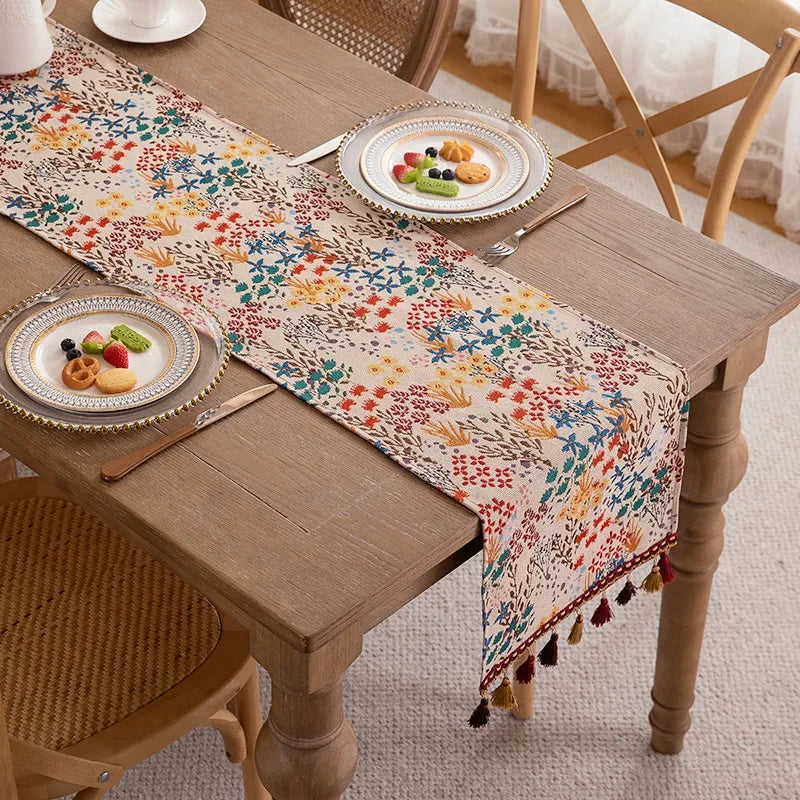 Chemin de table aux motifs floraux multicolores avec des glands décoratifs, sur une table en bois rustique avec des assiettes de fruits.