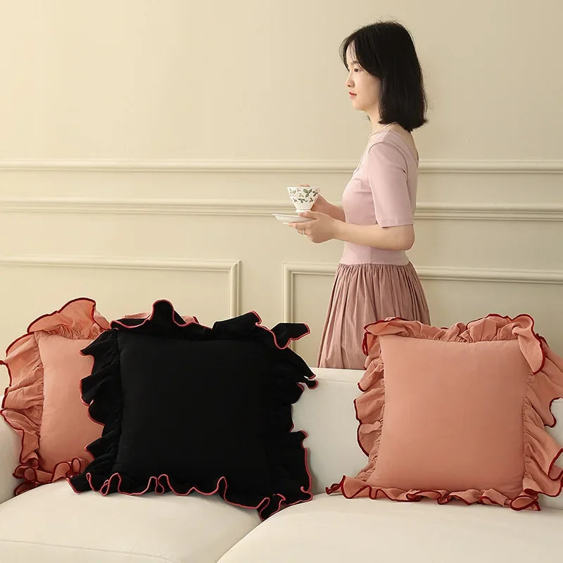 Une jeune femme élégante dans une tenue rose pastel se tient debout devant un canapé décoré de coussins rouges et noirs.