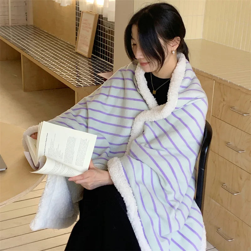 Une jeune femme assise dans un espace intérieur, enroulée dans une couverture à rayures blanches et violettes, lisant un livre.