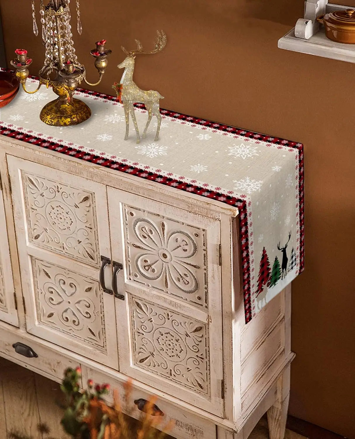 L'image montre un meuble de style rustique décoré de motifs fleuris sculptés, sur lequel sont posés des objets de décoration de Noël, comme un cerf lumineux et des chandeliers. Le meuble est recouvert d'une nappe ornée de flocons de neige.
