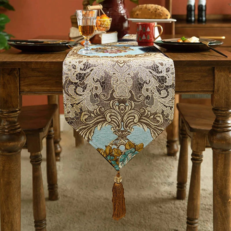 Une élégante chemin de table brodé, avec de belles motifs floraux et des glands, sur une table en bois rustique avec des éléments de décoration comme un verre de vin et une tasse.