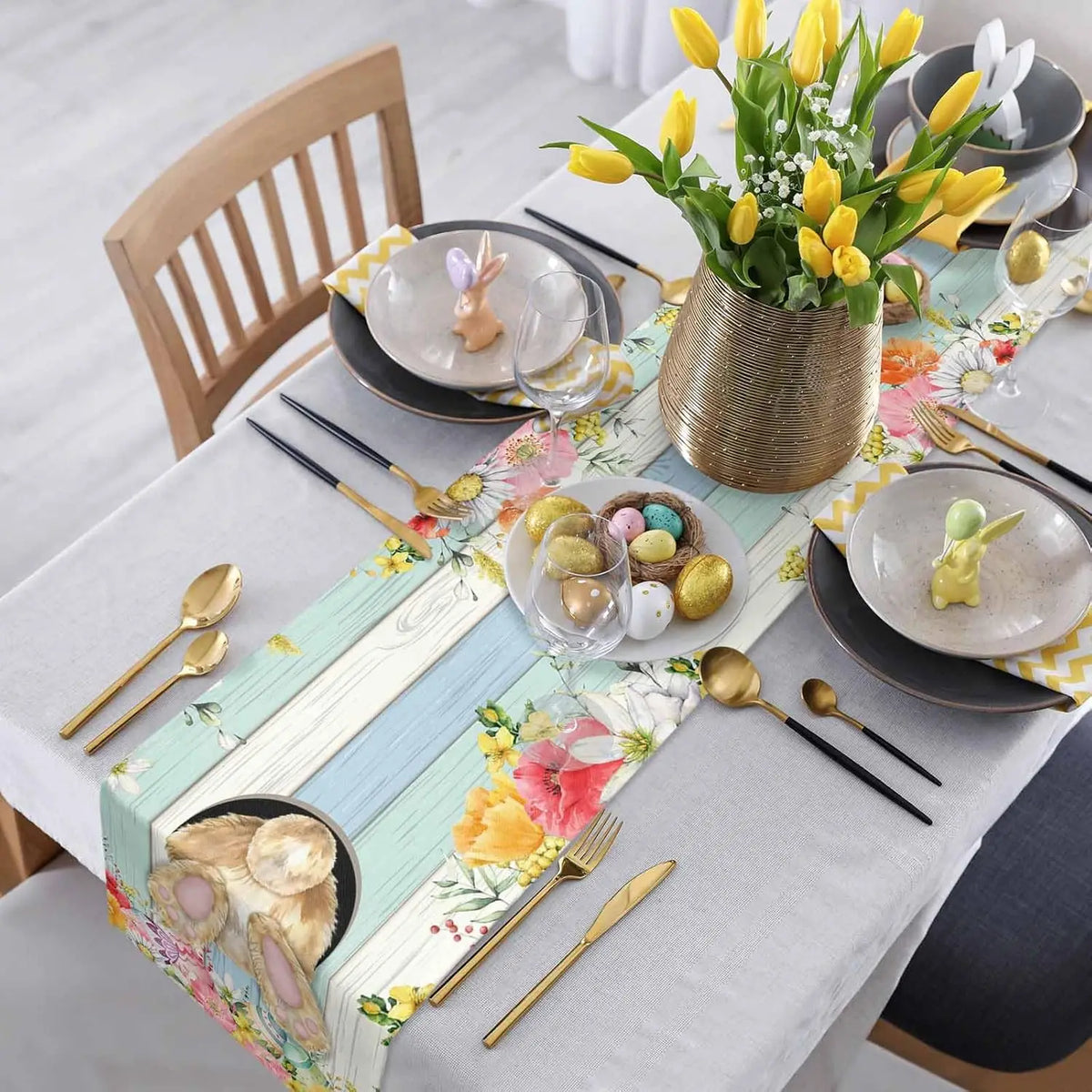 Une table festive décorée avec des fleurs de tulipes jaunes, des œufs colorés et des accessoires dorés, créant une atmosphère printanière et élégante.