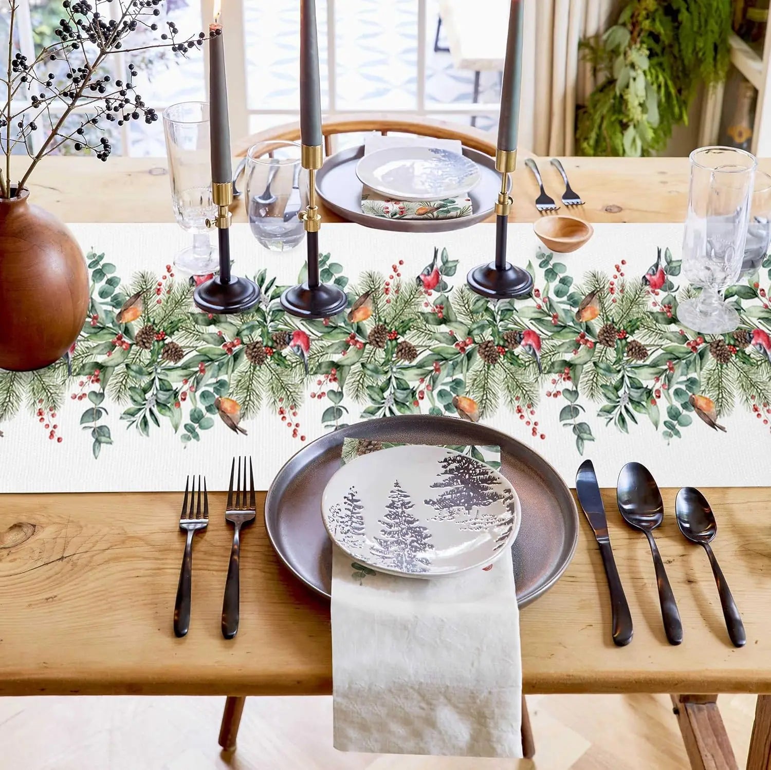 Cette belle table de Noël présente une décoration festive avec des branches de sapin, des baies rouges et des chandelles noires, le tout disposé sur un motif de feuilles vertes. Cette composition chaleureuse et élégante crée une ambiance propice à un repas de fête.