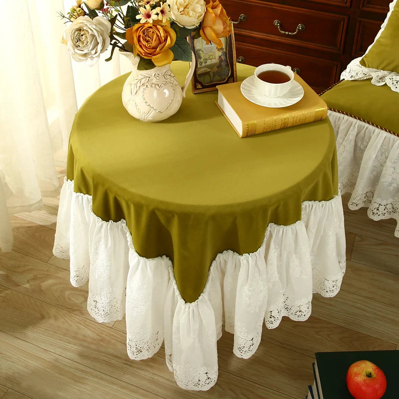 Une table ronde avec une nappe vert olive ornée de dentelle blanche, sur laquelle sont posés un bouquet de fleurs dans un vase, un livre ancien et une tasse de thé, créant une atmosphère chaleureuse et élégante.