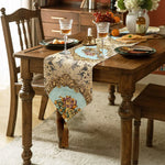 Une table à dîner rustique avec un chemin de table décoré de motifs floraux et de franges dorées, entouré de délicieux plats et verres de vin.