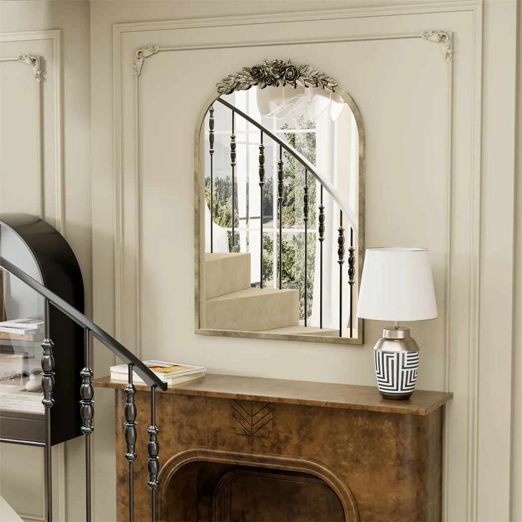 Un beau miroir ouvragé sur le mur, surmonté d'un joli ornement floral, reflétant une fenêtre donnant sur un paysage verdoyant. Une console en bois accueille une lampe de style élégant et une décoration aux motifs géométriques.