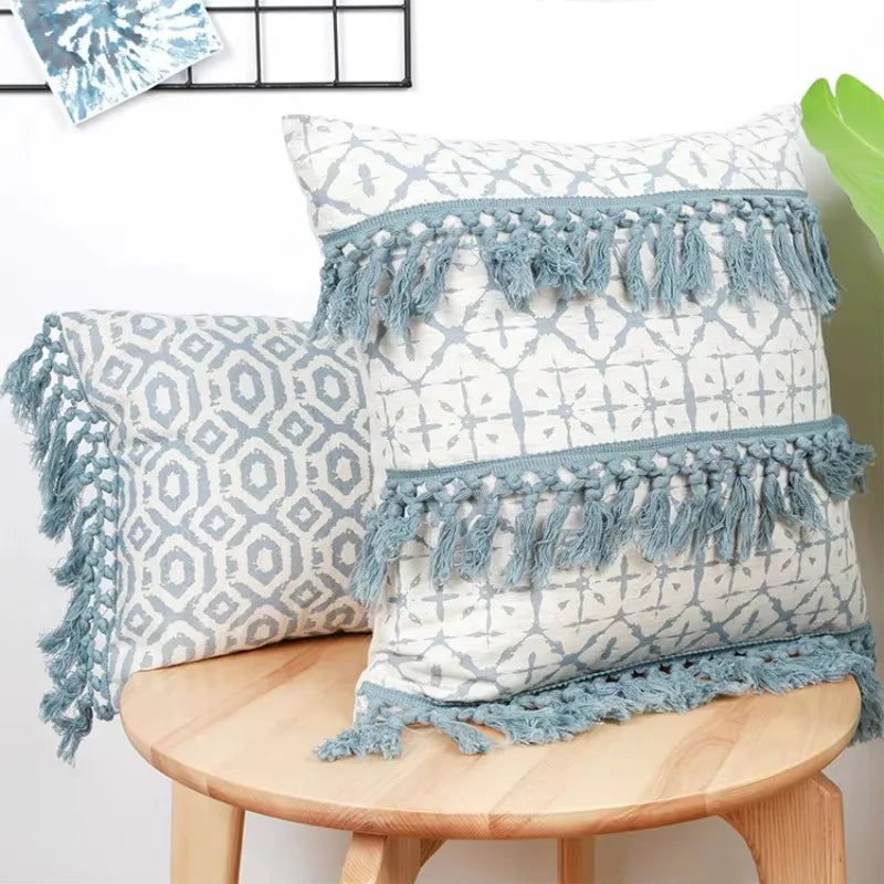 Deux coussins décoratifs en tissu blanc et bleu orné de franges et de motifs géométriques posés sur un tabouret en bois.