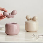 Deux jolies boîtes de stockage en céramique blanche et rose pastel, avec des formes douces et des couvercles en forme de fleurs, créant une atmosphère élégante et apaisante sur une surface blanche.