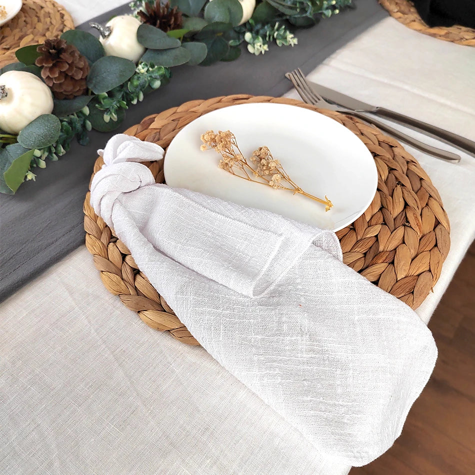 Un beau service de table avec une assiette blanche, des accessoires décoratifs et une nappe en lin blanc sur un plateau tressé.