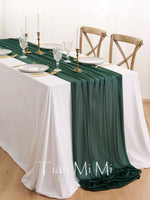 Cette image montre une table dressée avec une nappe blanche et un chemin de table vert foncé. Sur la table, on peut voir des assiettes, des verres, des chandelles et des couverts. Les chaises sont en bois naturel avec un dossier croisé.