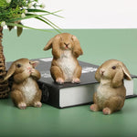 Une famille adorable de lapins en céramique, posée sur un livre avec des plantes verdoyantes en arrière-plan.