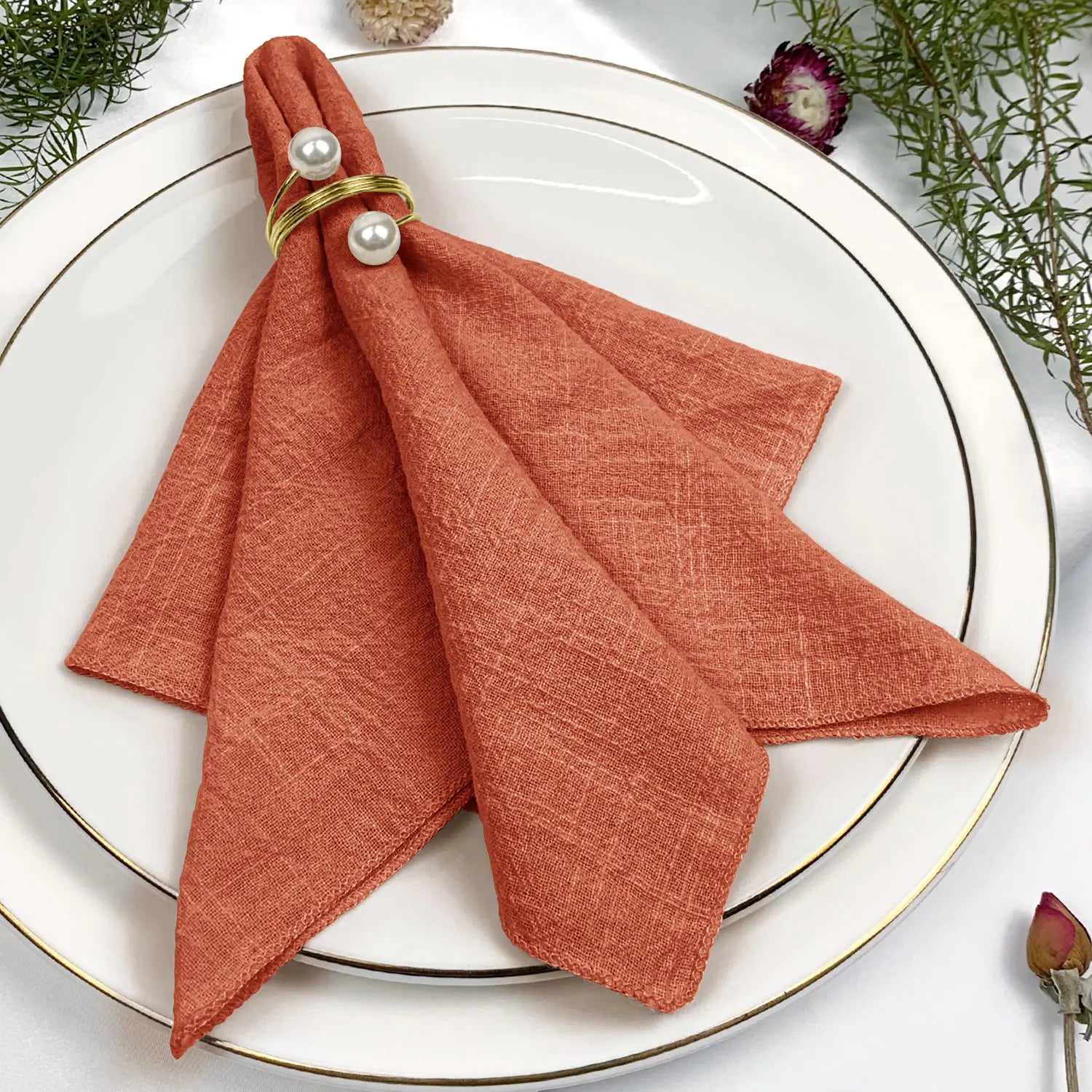 Serviette en lin de couleur orange disposée sur une assiette blanche, agrémentée de perles blanches sur un ruban doré.