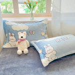 Coussin décoratif bleu clair avec des lapins en peluche blancs et une enseigne "Happy" sur le lit d'un salon, avec des fleurs dans un vase en arrière-plan.