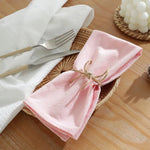 Serviette de table rose, couteau et fourchette posés sur un panier en osier, sur un plan de travail en bois clair, avec des fleurs blanches en arrière-plan.