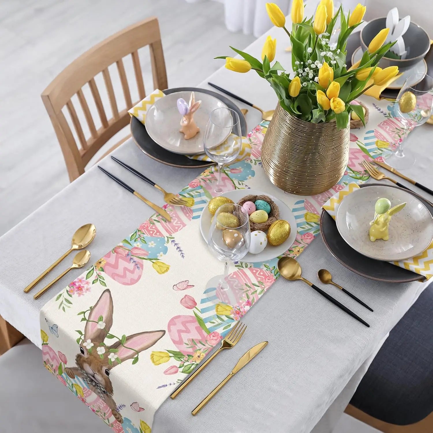 Une table de fête printanière avec des tulipes jaunes dans un vase doré, des assiettes et des couverts dorés, des oeufs de Pâques colorés, et une nappe avec des motifs floraux et de lapins.