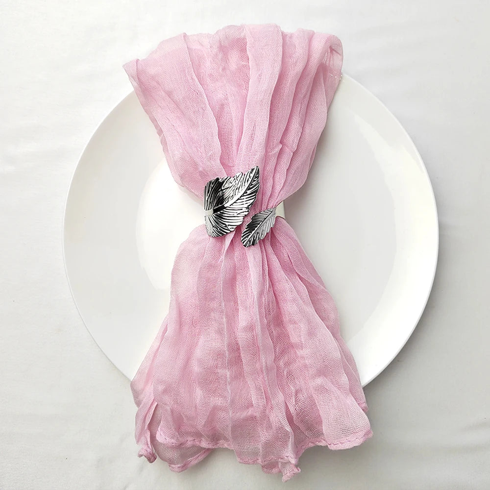 Un foulard rose poudré, souple et élégant, noué par une broche décorative en forme de plumes, se dresse sur un plat blanc.