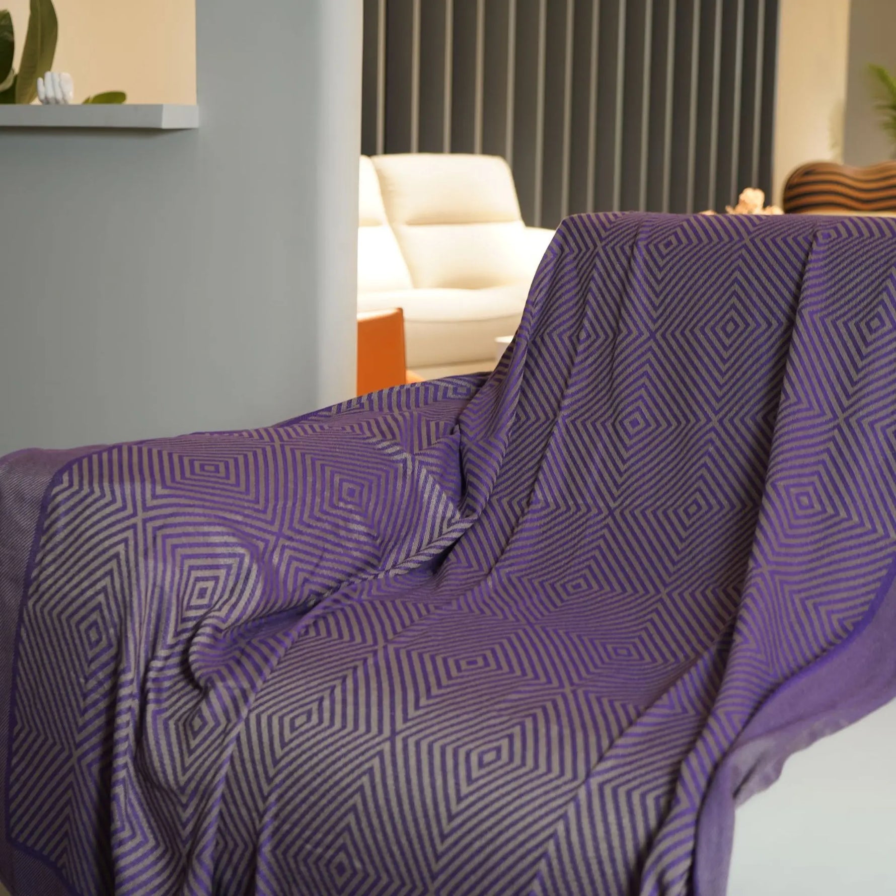 Couverture rectangulaire en coton de couleur violette et grise aux motifs géométriques décorée dans un salon avec des meubles beiges.