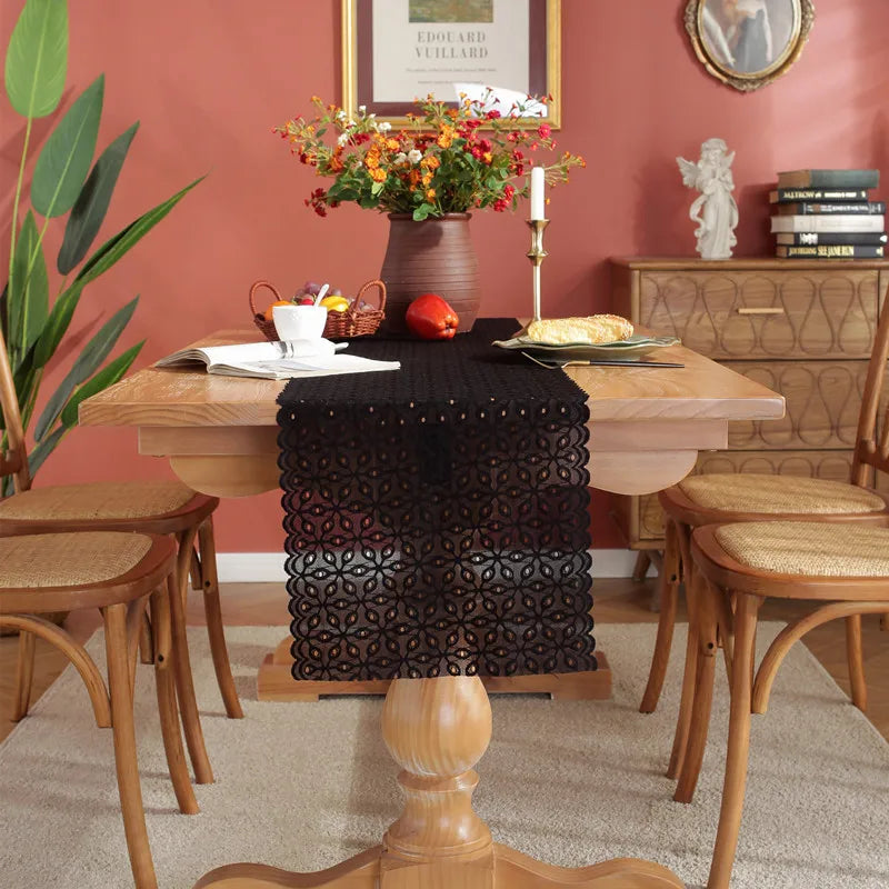 Une scène élégante de salle à manger avec une table en bois, un bouquet de fleurs colorées, des chaises en rotin, et un chemin de table décoratif noir en dentelle.