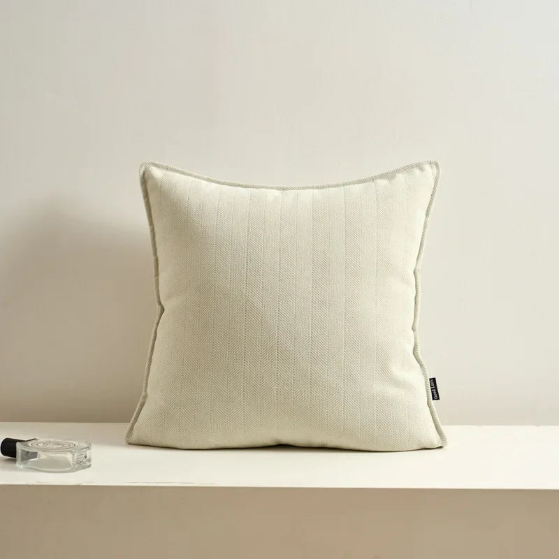 Coussin carré blanc cassé avec bordure décorative, posé sur une surface blanche avec deux accessoires.