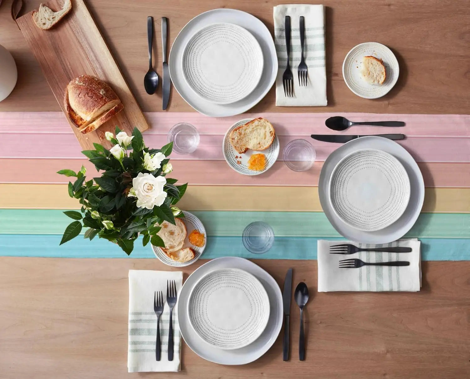 Sur une table en bois, une mise en place de table soignée avec des assiettes et des couverts blancs, des fleurs et du pain, suggérant un repas convivial et harmonieux.